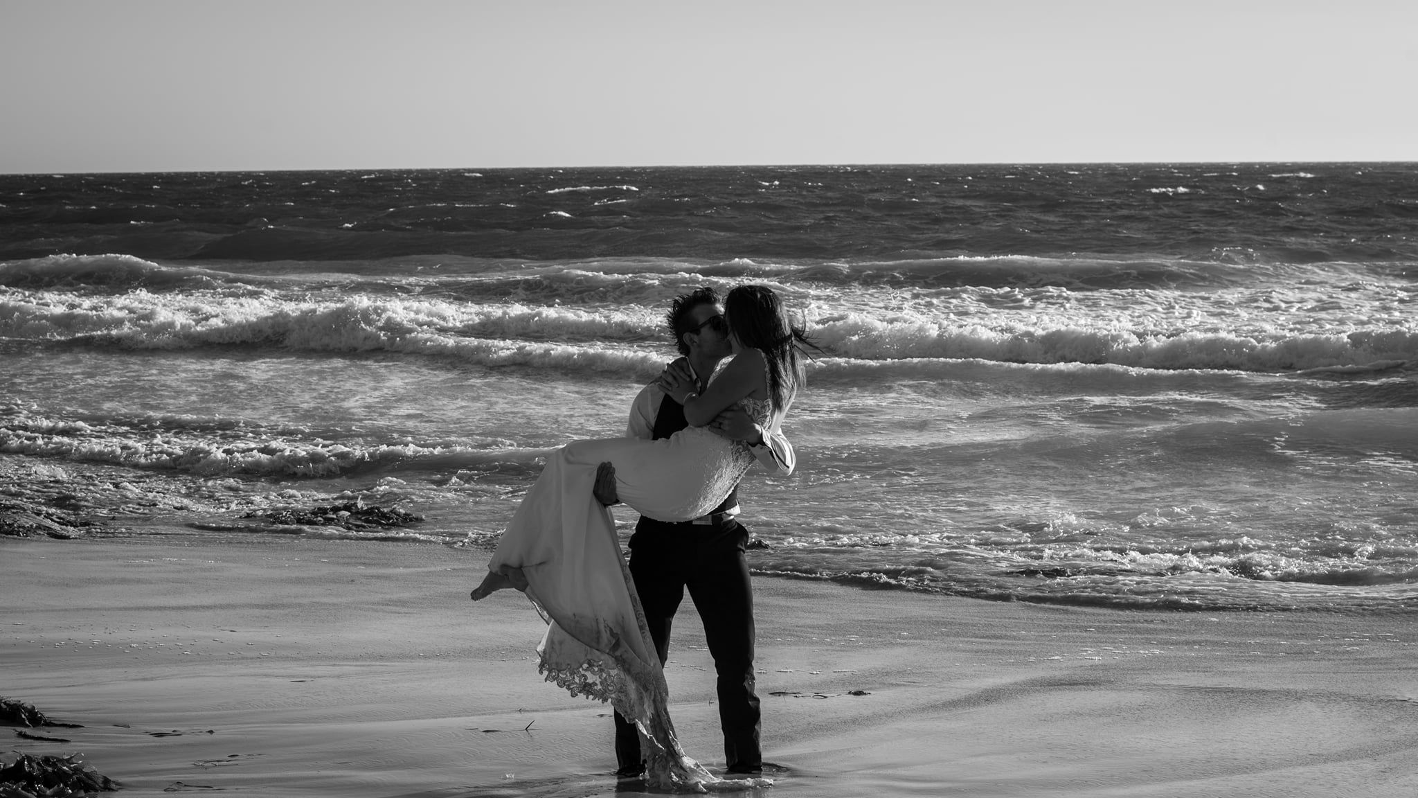 A silhouetted couple embracing on a beach at sunset with waves crashing behind them.