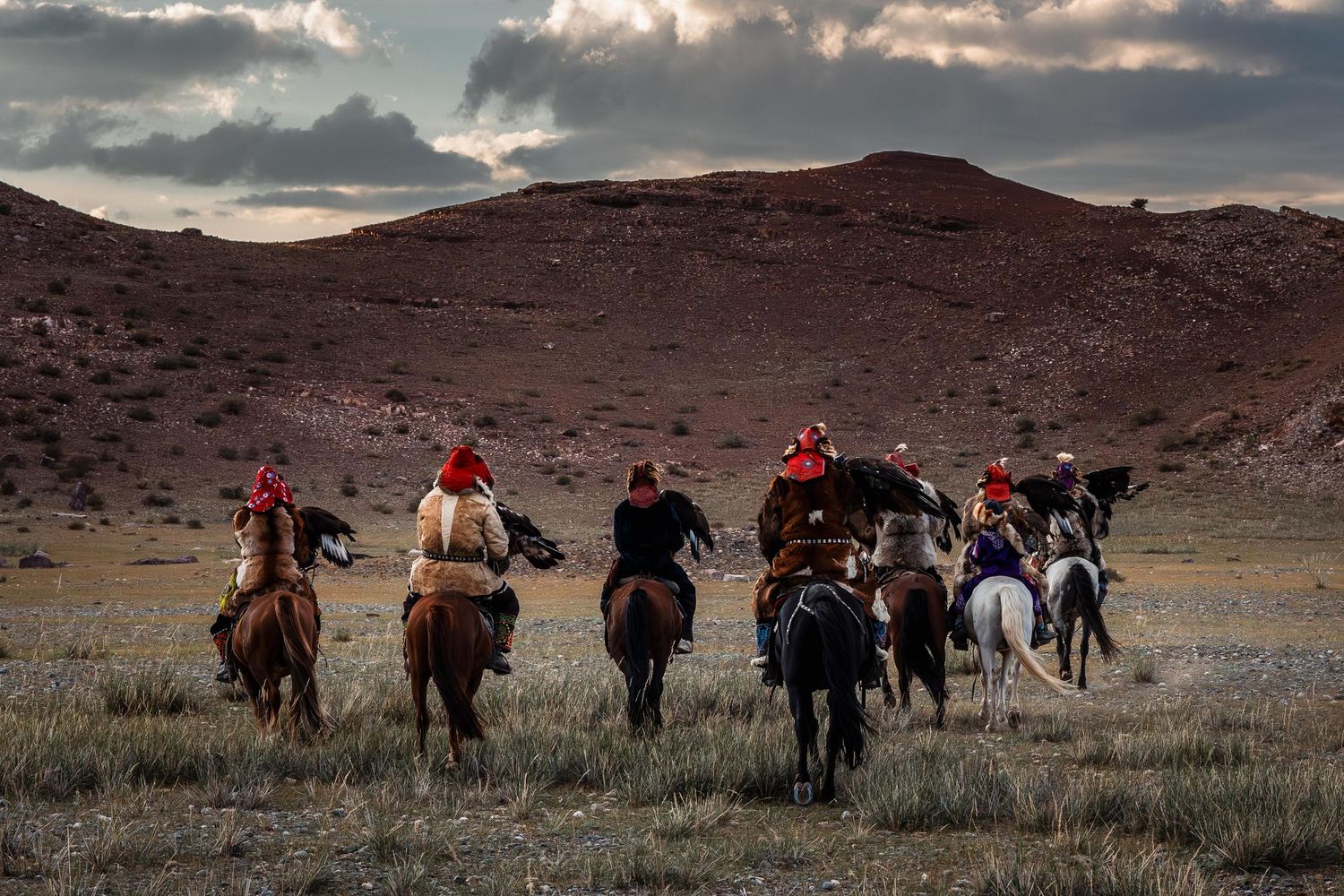Group of horseback riders traverse rugged desert terrain under dramatic cloudy sky.