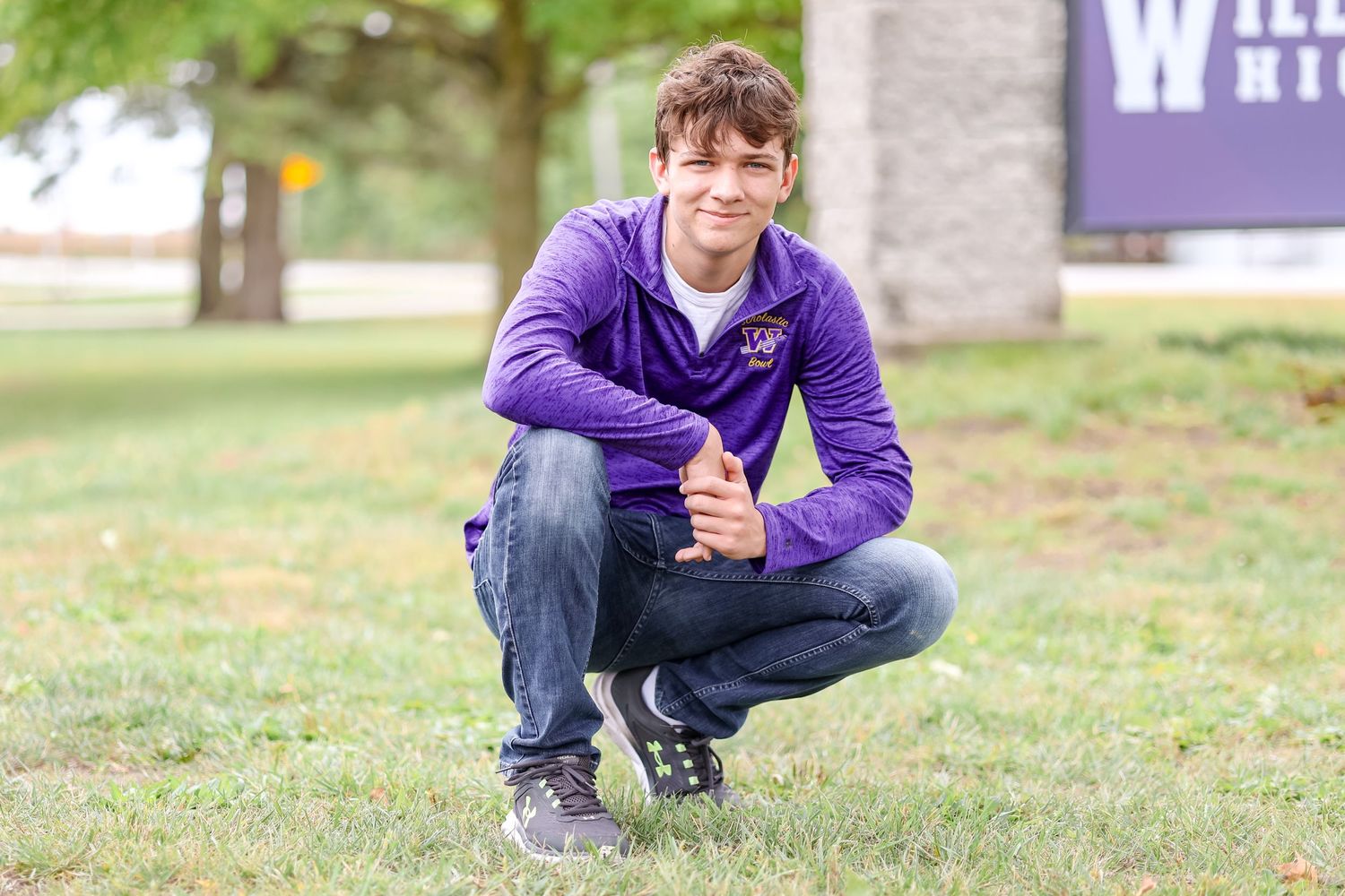 Senior in purple pullover squats down in grass with school sign in background