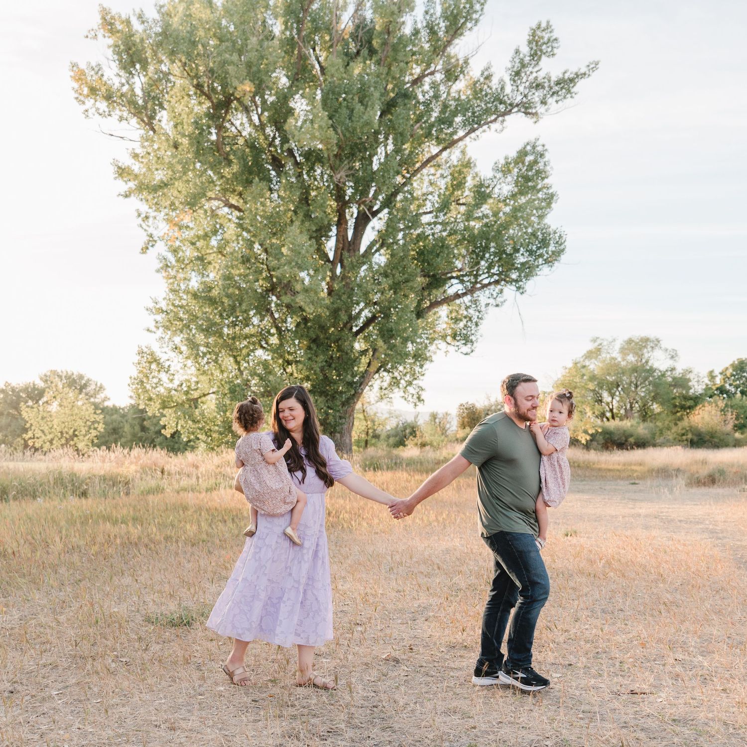 Playful family moment captured in a sun-drenched field with tall trees.