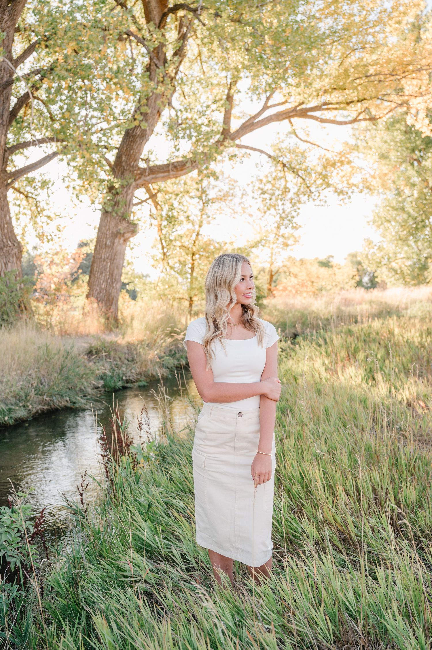 Woman in white dress standing by stream in tall grass during golden sunset.