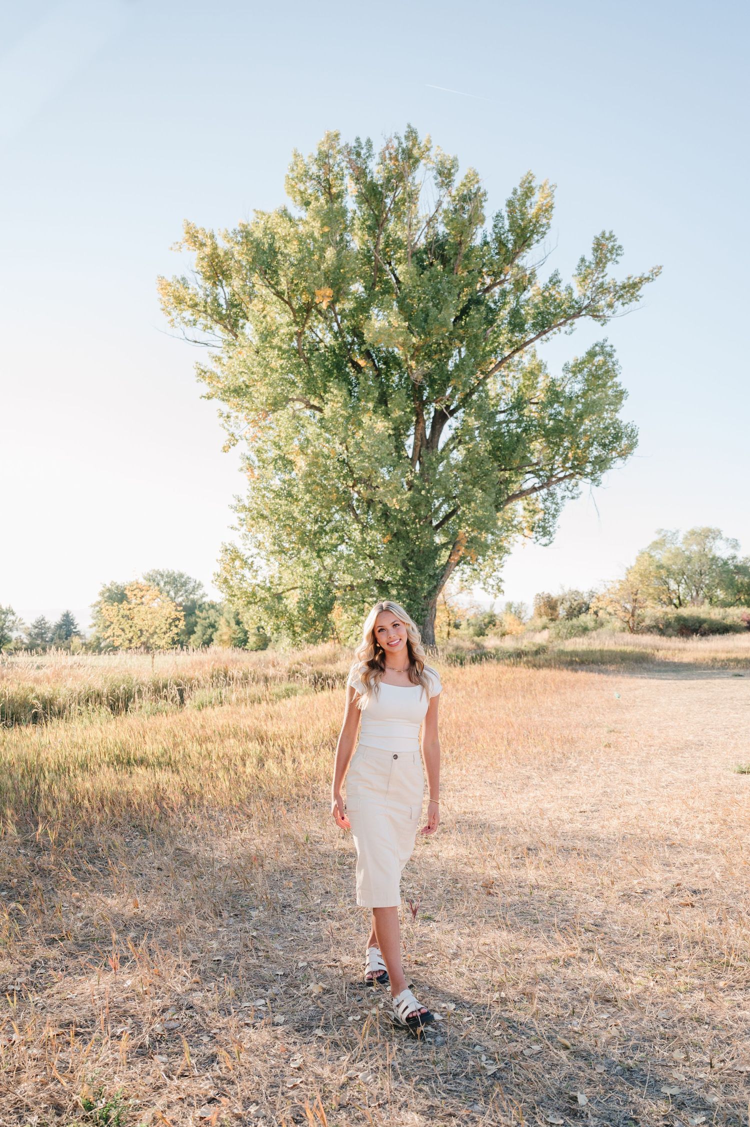 Woman in white dress poses near tall tree in rustic outdoor setting.