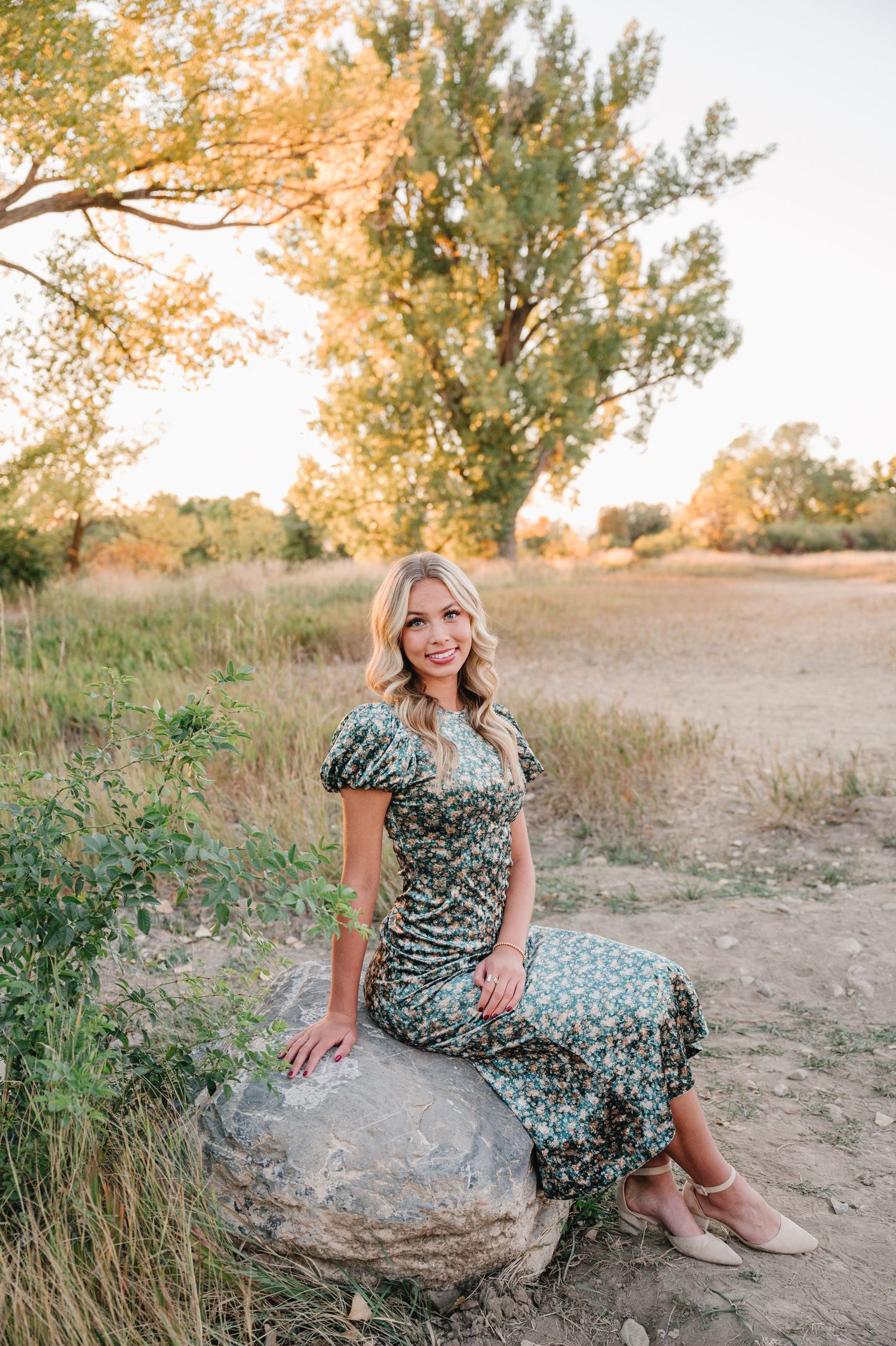 Woman in floral dress sits on large rock during sunset photoshoot.