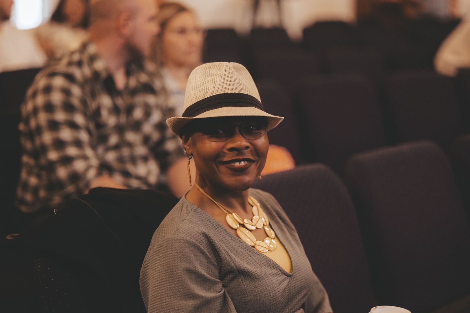 Person wearing a hat and necklace smiling during an indoor event.