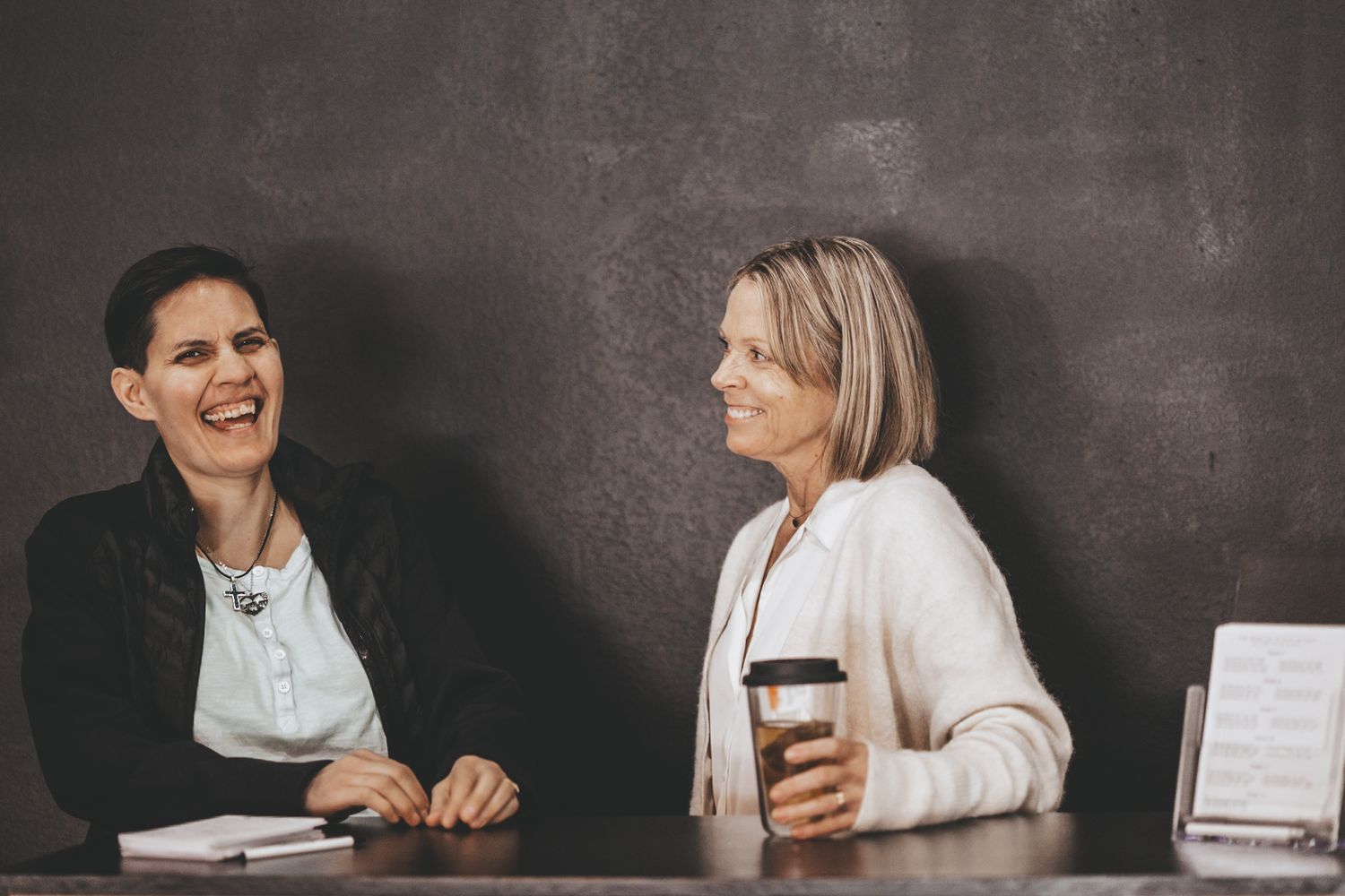 Two people laughing during a casual meeting with coffee against dark wall.