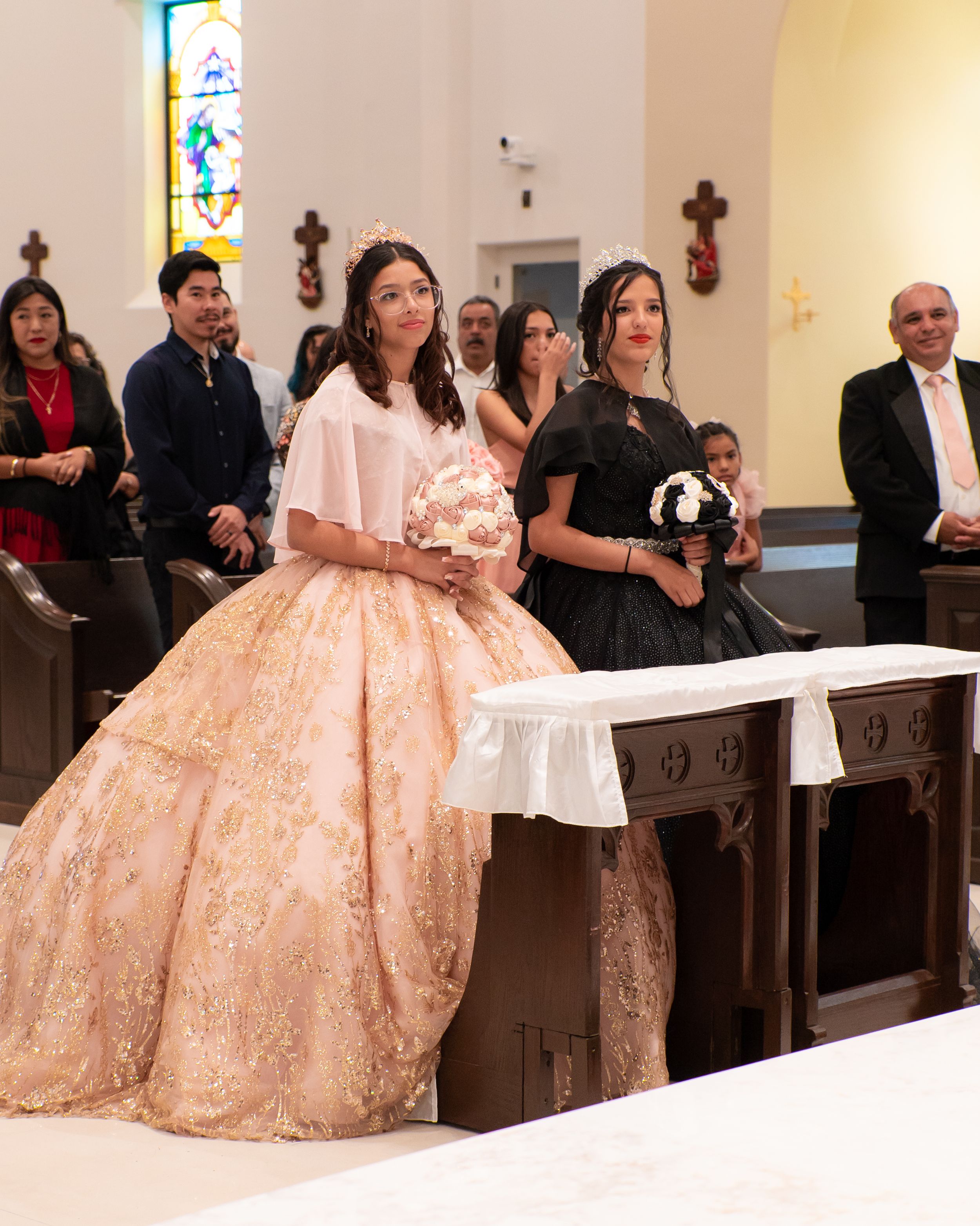 Young woman in ornate gold quinceañera gown walking down church aisle with escort in black dress.