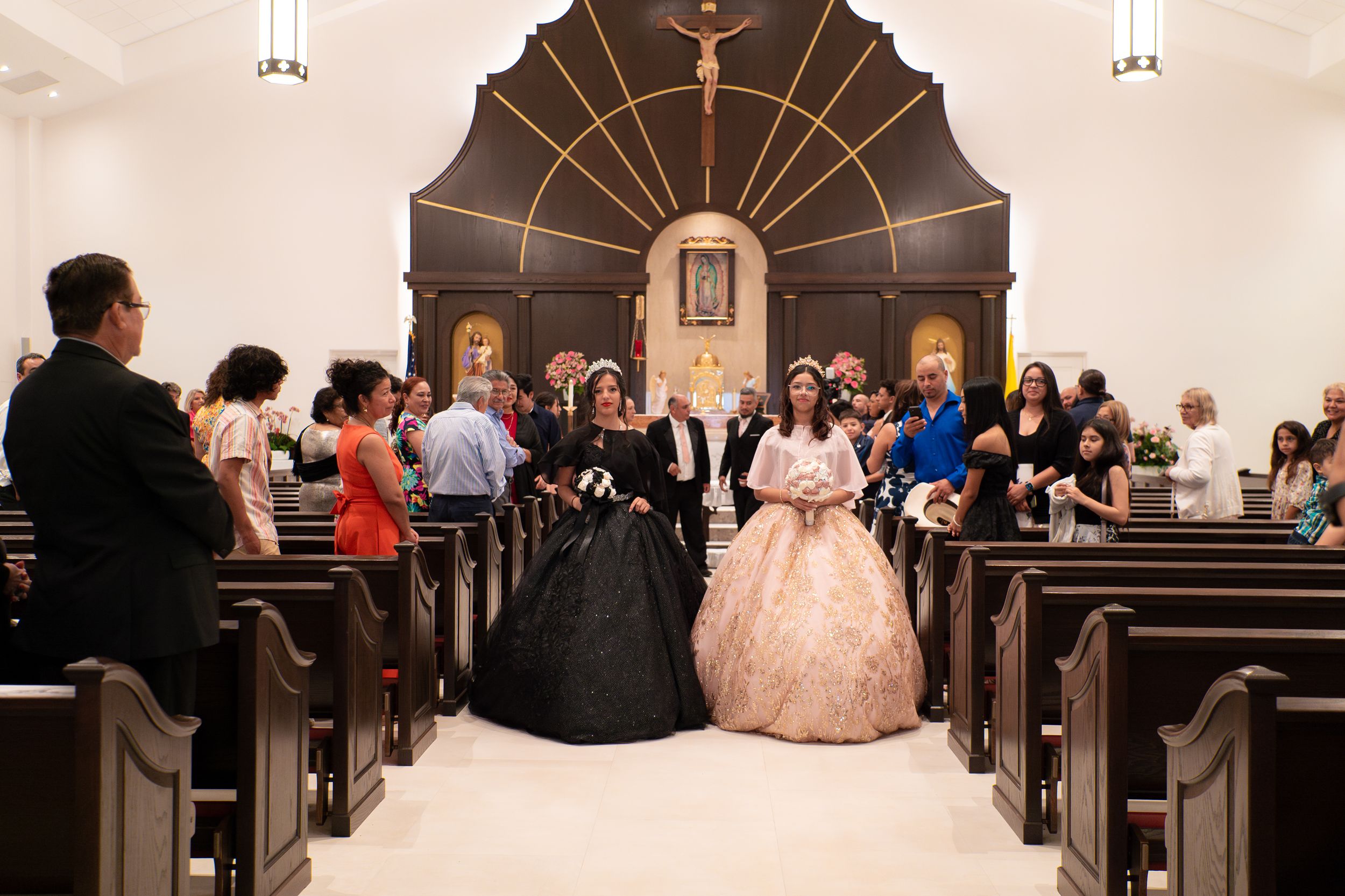 Two girls in stunning black and gold quinceañera ball gowns walking down a church aisle together.