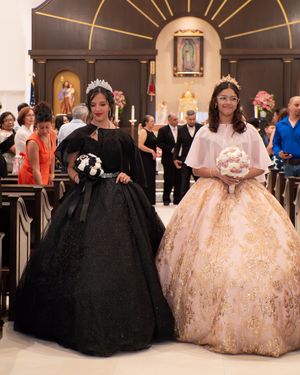 Two quinceañera celebrants in black lace and gold embroidered ball gowns posing in a church.