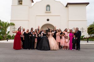 Large quinceañera family group photo posing outside a white Spanish-style church after the ceremony.