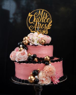 Two-tier pink quinceañera cake with gold Mis Quince Anos topper, black and gold ball decorations.
