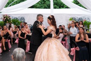 Young woman in pink quinceañera gown shares first dance with father under white tent at celebration.
