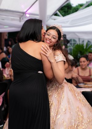 A young Quinceañera in a pink sequin gown shares a warm hug with a woman in a black dress.