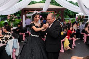 A Quinceañera in a black ball gown dances with an older man at an outdoor celebration.