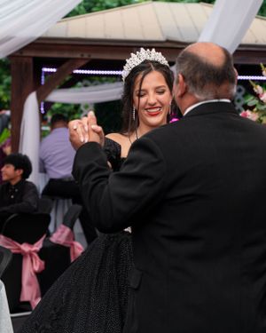 A smiling young woman in a black gown and tiara shares a dance with an older man at her Quinceañera.