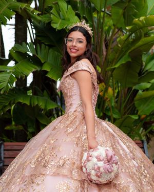 Young woman in rose gold quinceañera gown with crown and bouquet posing among tropical green foliage.