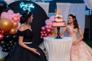 Two girls in formal gowns admire a pink Quinceañera cake at a party with balloon decorations.