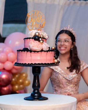 A smiling teen in a rose gold sequin dress poses beside her pink two-tier Quinceañera cake.