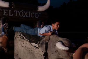 A young boy playfully climbs on a mechanical bull named El Toxico at a nighttime event.