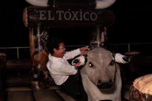 A person interacts with a mechanical bull named El Toxico at an outdoor nighttime attraction.