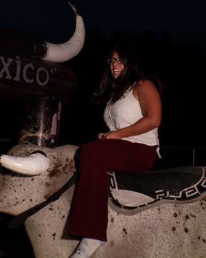 A young woman smiles while sitting on a mechanical bull named El Toxico at a nighttime event.