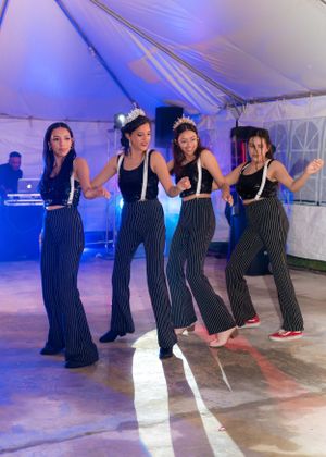 Four young women in matching pinstripe outfits and tiaras perform a choreographed dance at a Quinceañera.