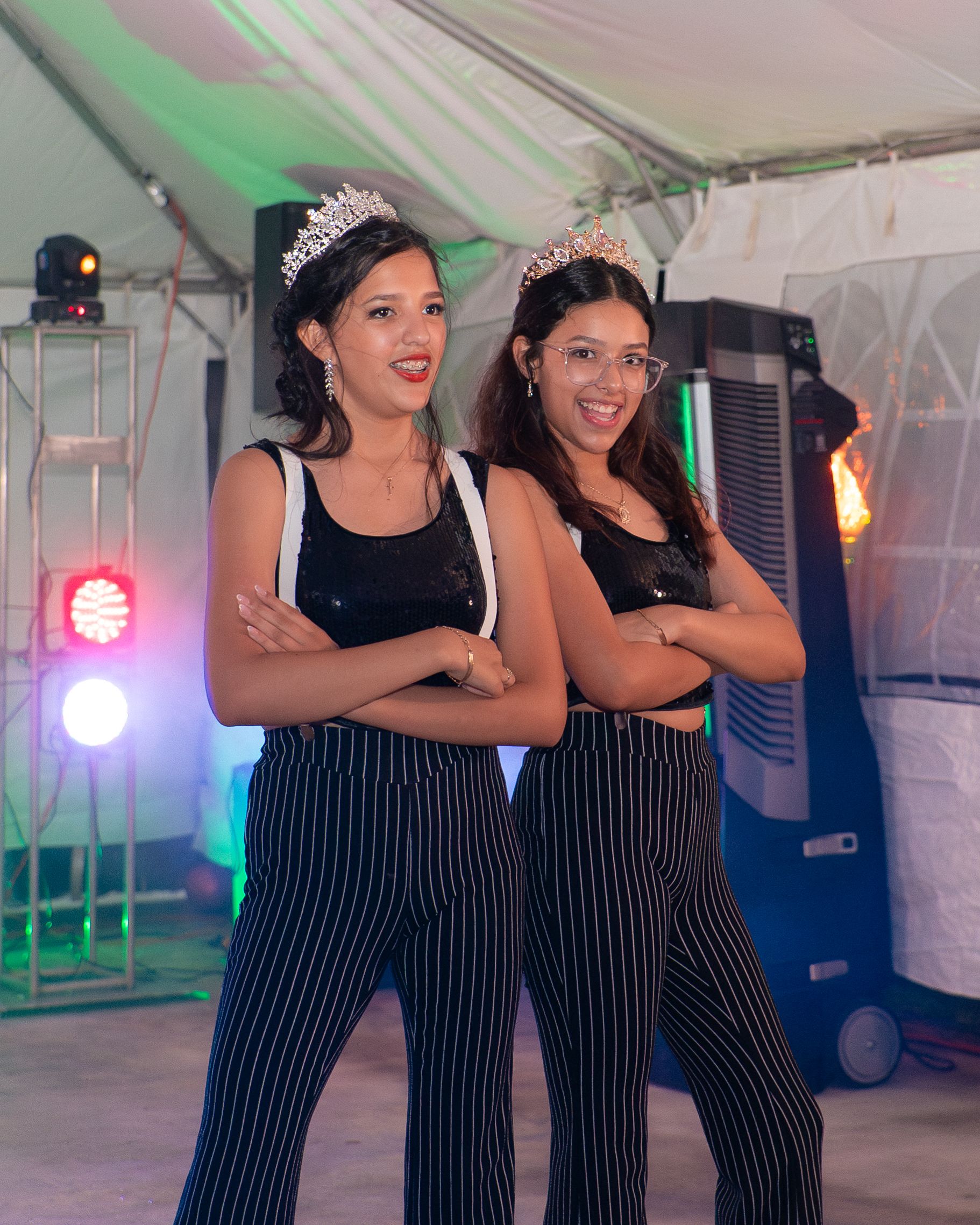 Two women in matching black striped outfits and tiaras pose with crossed arms at a festive party event.