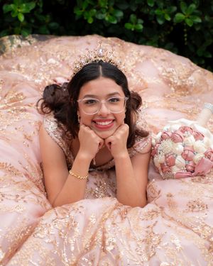 Smiling quinceañera in rose gold embroidered gown and crown lying down holding a floral bouquet outdoors.