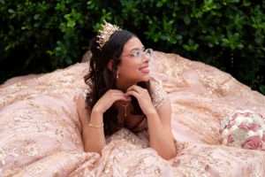Quinceañera in rose gold ball gown with tiara and glasses posing dreamily against lush green hedge backdrop.