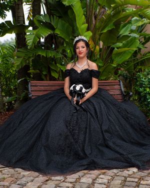 Quinceañera in elegant black ball gown with tiara seated on a bench surrounded by tropical plants.