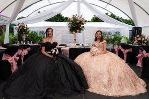 Two quinceañeras in stunning black and rose gold ball gowns seated at decorated tables under a white tent.