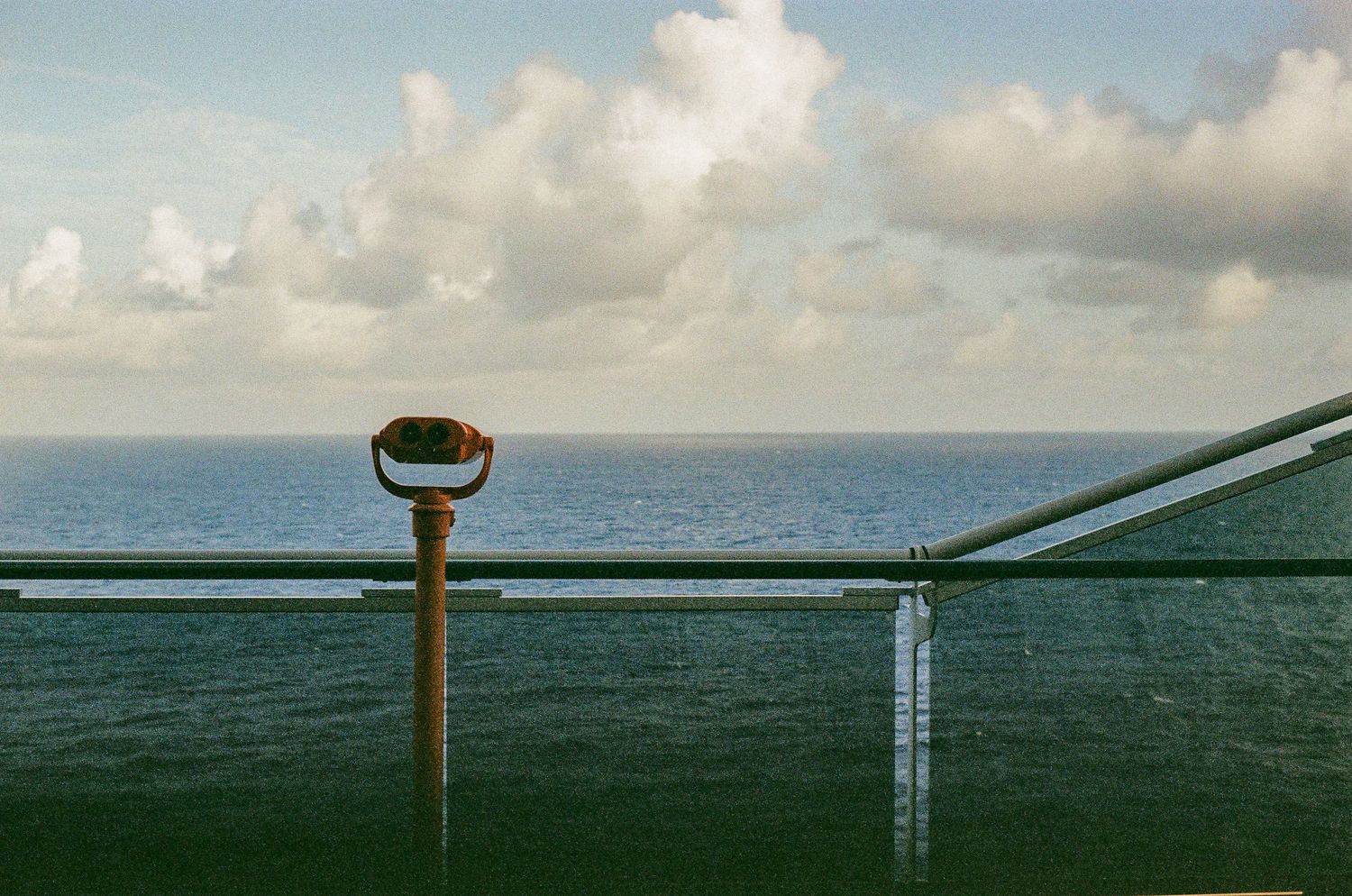 Coin-operated viewing binoculars mounted on railing overlooking ocean horizon under cloudy sky.