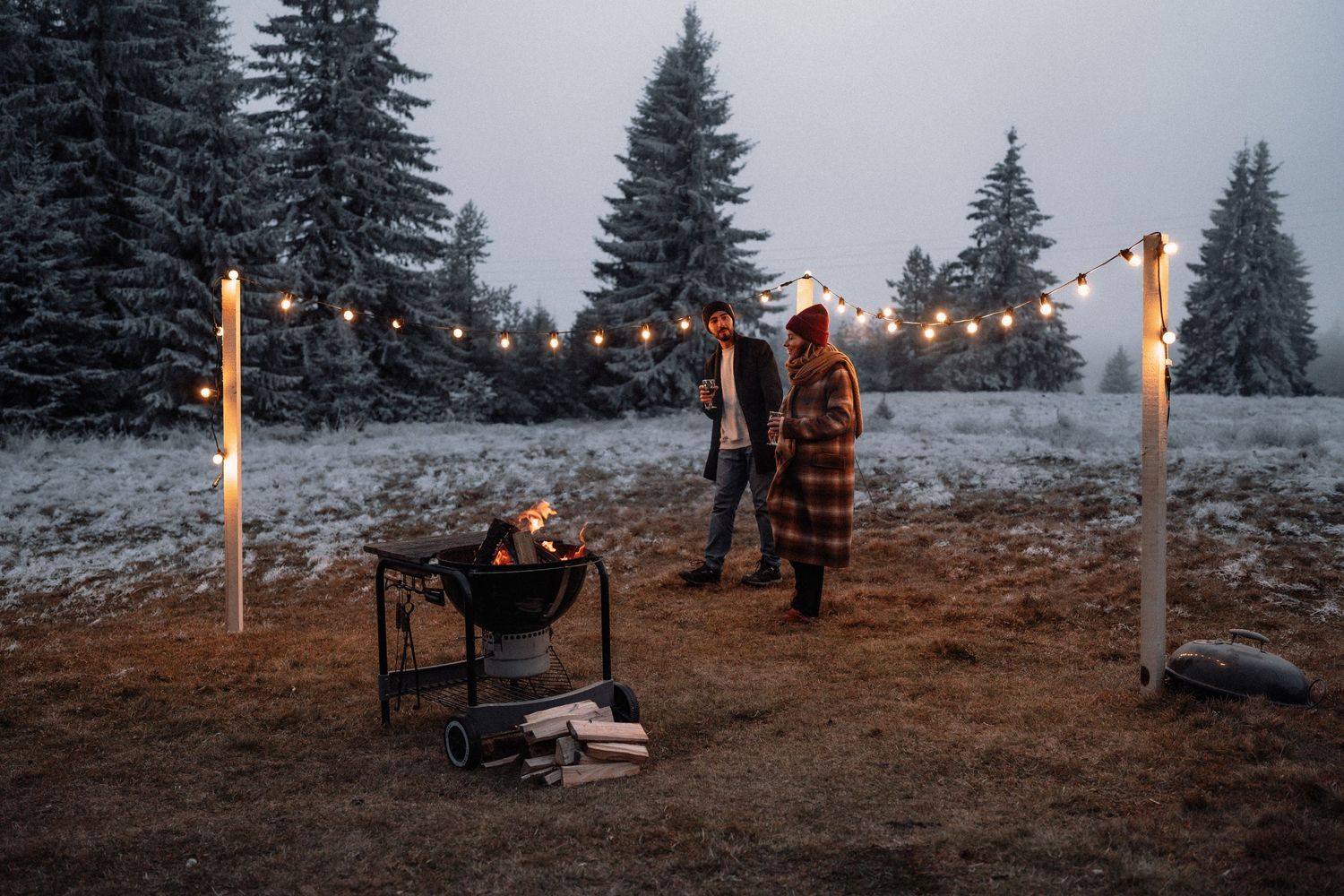 A winter wedding celebration at dusk with string lights illuminating snow-covered evergreen trees in the background.