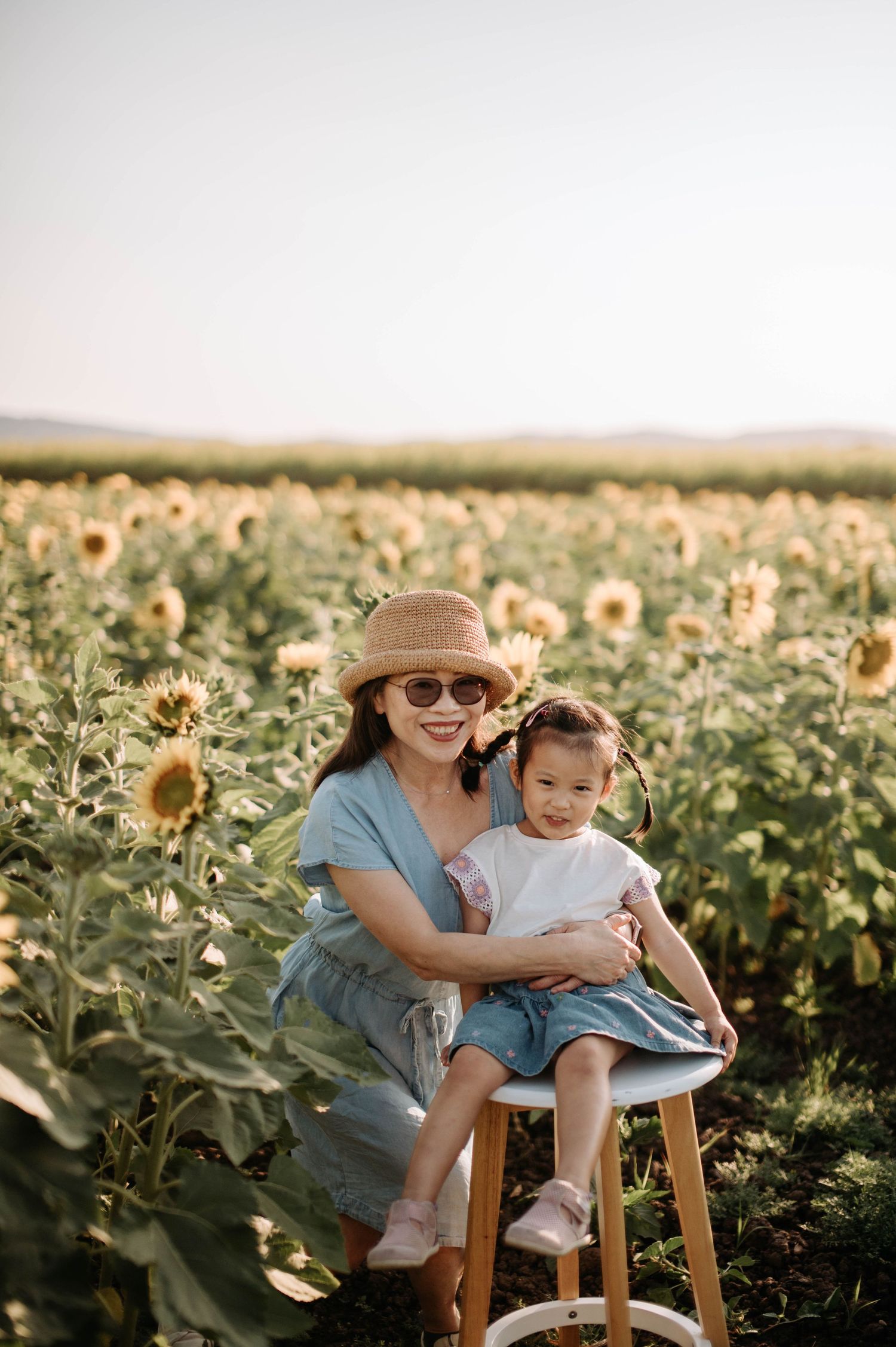 Sitting on a wooden stool in a sunflower field wearing a summer dress and hat.
