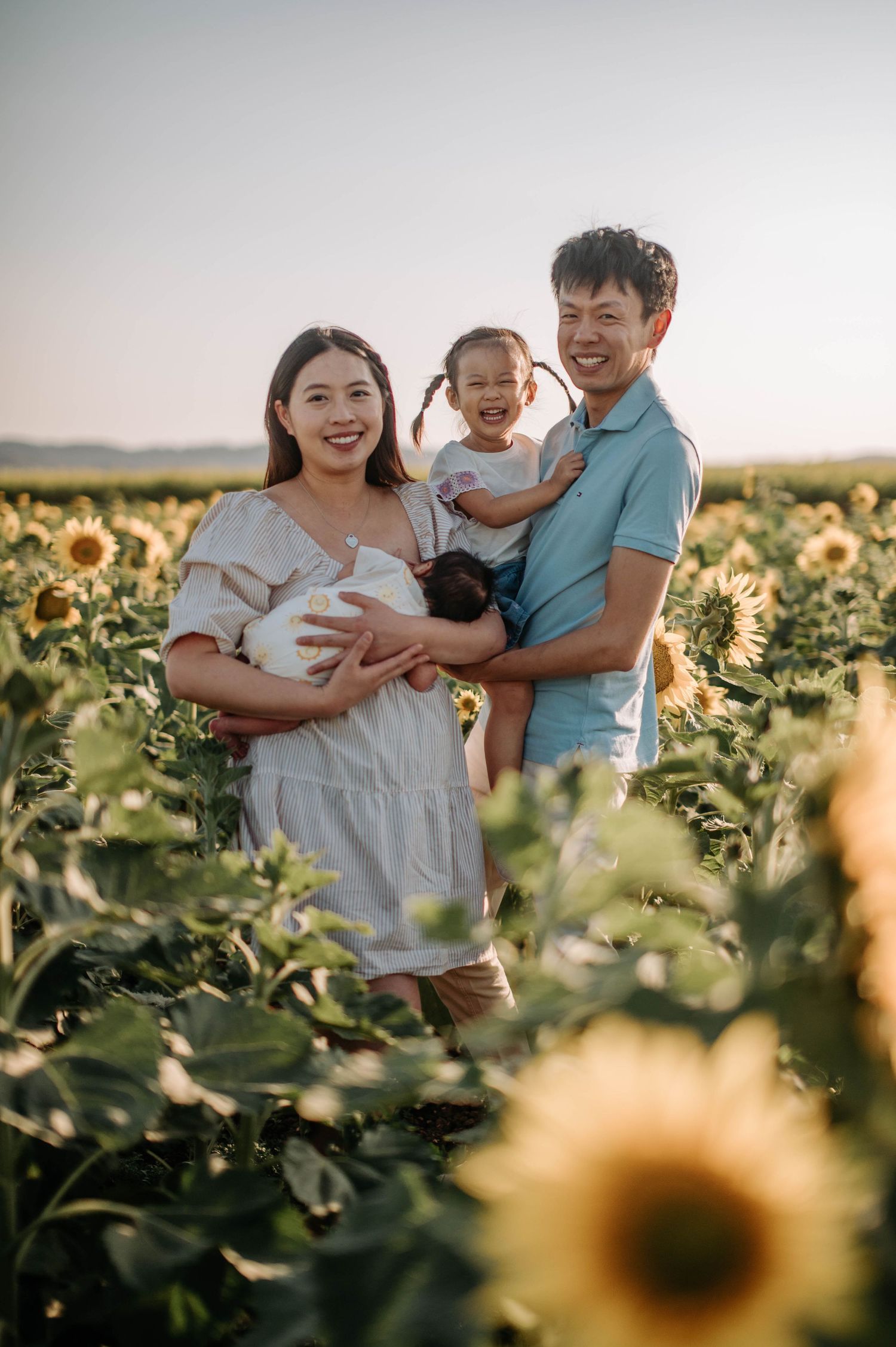 Family of three laughing together in sunlit field of blooming sunflowers.