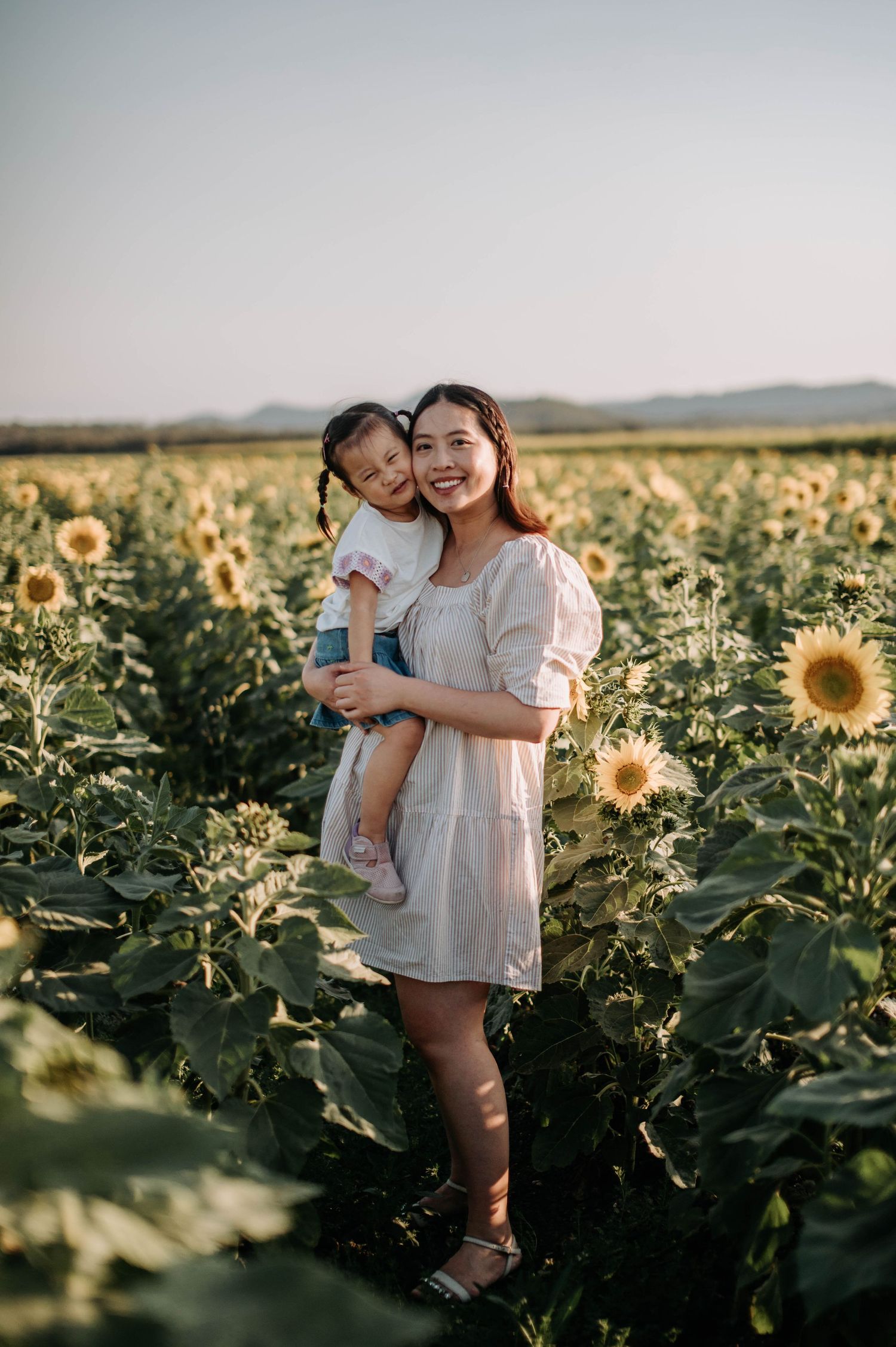 Mother holds child while standing amidst blooming sunflowers during sunset.