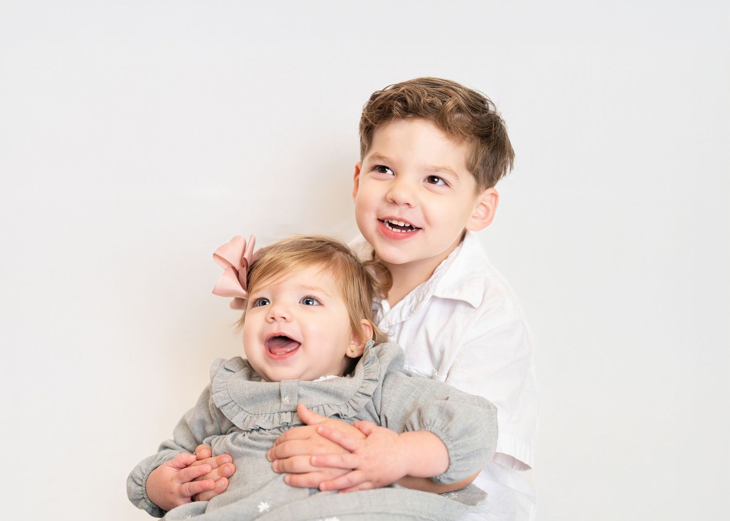 Two young children in white and gray outfits share a joyful moment against a light background.
