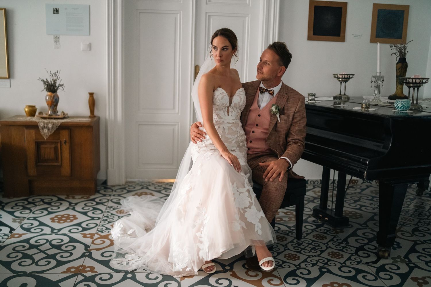 Bride in lace gown and groom in tan suit pose elegantly by a grand piano on ornate tiled floors.