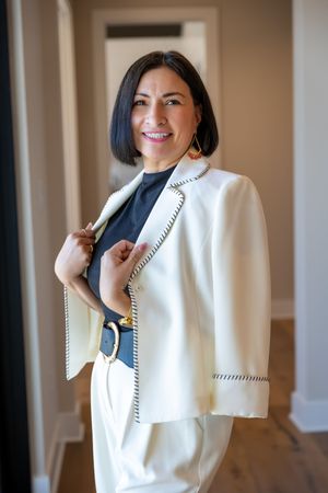 Woman in white blazer and pants suit with black top and belt, smiling indoors.