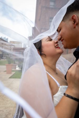 A joyful bride and groom share a romantic moment under a flowing white veil outdoors on their wedding day.