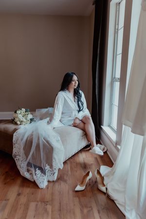 Bride in white robe sitting by window with lace veil, bridal bouquet, and white heels on hardwood floor.