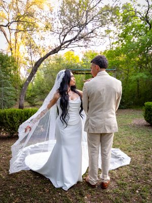 Bride in white gown and lace veil stands with groom in tan suit, backs turned, in a lush garden setting.