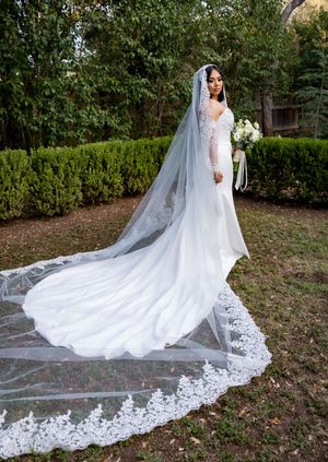 Bride in elegant long-sleeve lace wedding gown with cathedral veil and bouquet, posing outdoors in garden setting.