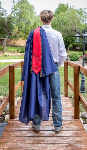 Graduate in blue gown and red honor stole walks across wooden bridge, viewed from behind.