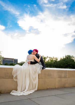 A couple with blue and pink hair sharing a romantic kiss on a rooftop in wedding attire under a blue sky.