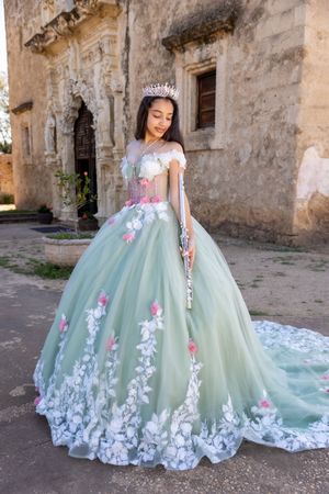 Young woman in mint green quinceañera ball gown with floral appliqués and tiara poses before historic stone building.