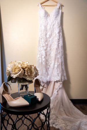 White lace wedding dress hanging on wall above table with nude heels, roses bouquet, and small teal jewelry box.