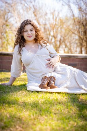Pregnant woman in white dress sitting on grass with tiny cowboy boots, smiling outdoors in natural light.