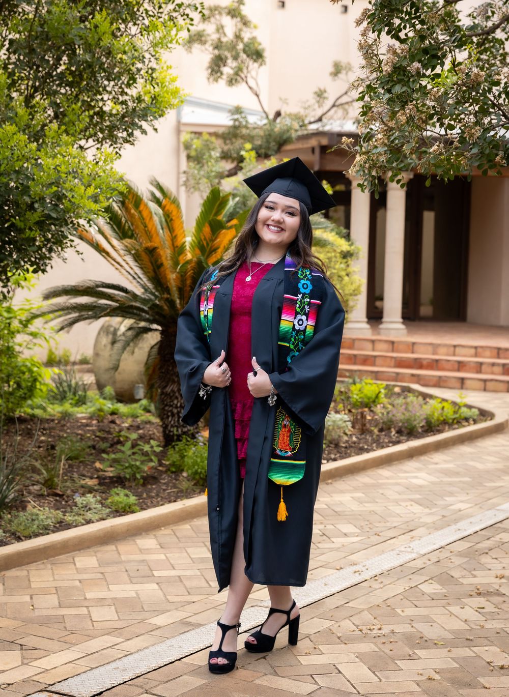 A smiling graduate in cap and gown with colorful stoles poses on a brick path surrounded by lush greenery.