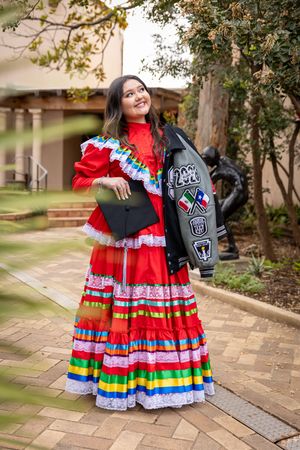 Young woman in colorful traditional Mexican dress and leather jacket holds graduation cap outdoors.