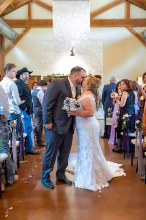 Bride and groom share first kiss at indoor rustic wedding chapel with guests standing in the aisle.